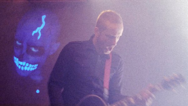 A guitarist is seen with a ghostly figure behind them.